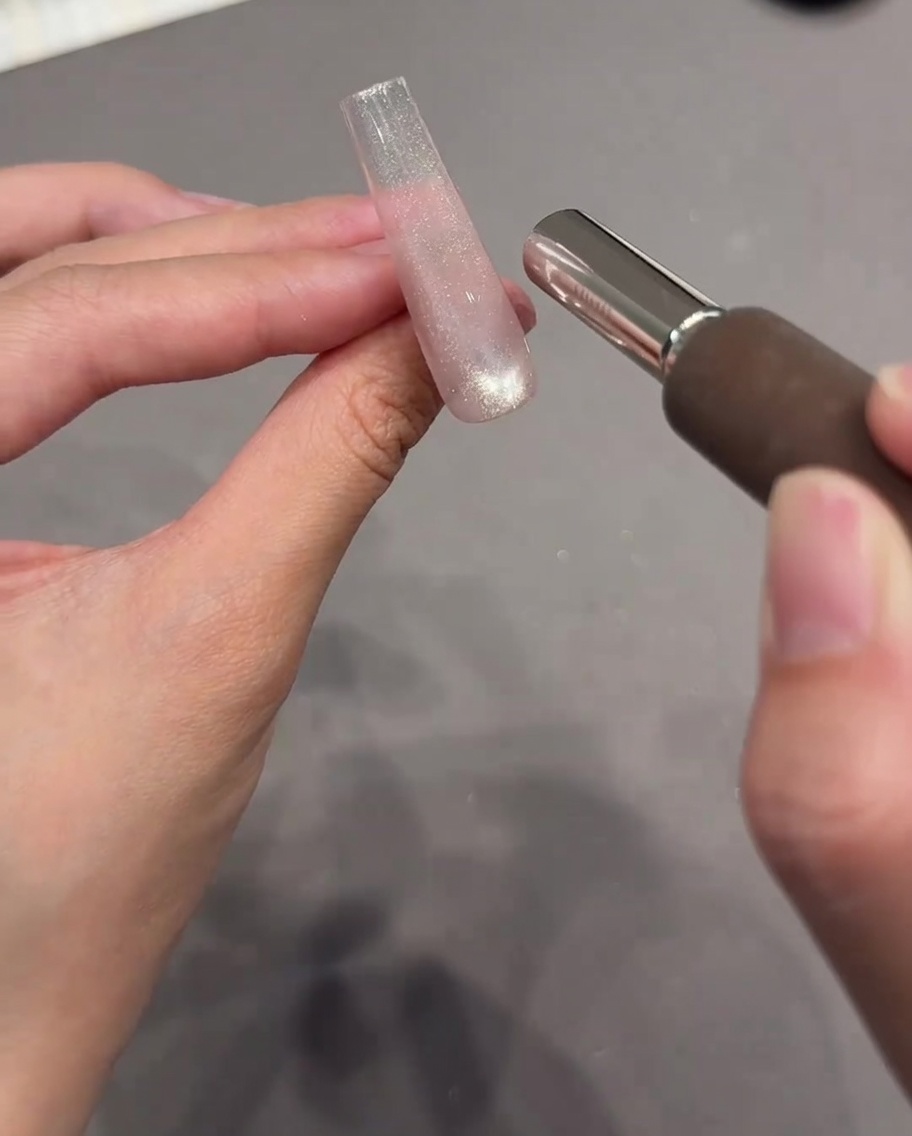Una mano che tiene un magnete cilindrico vicino a unestensione per unghie chiara rivestita di gel cat eye scintillante bagnato.