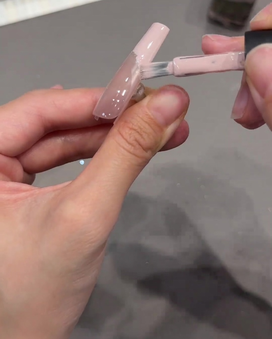 Un pennello per unghie che applica gel rosa nude opaco sulla punta di ununghia, usando uno sticker curvo trasparente come confine pulito.