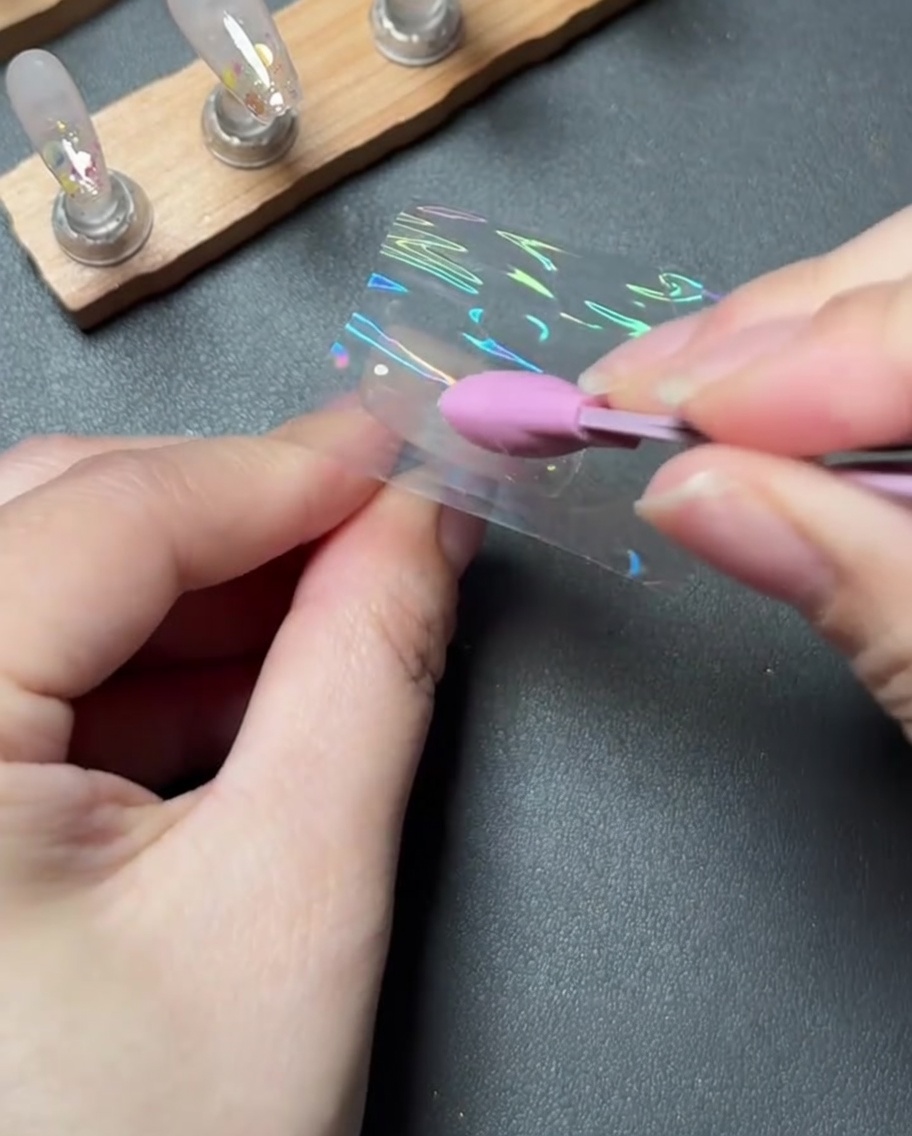 Uno strumento in silicone rosa che preme e leviga saldamente un pezzo di pellicola transfer olografica su ununghia.