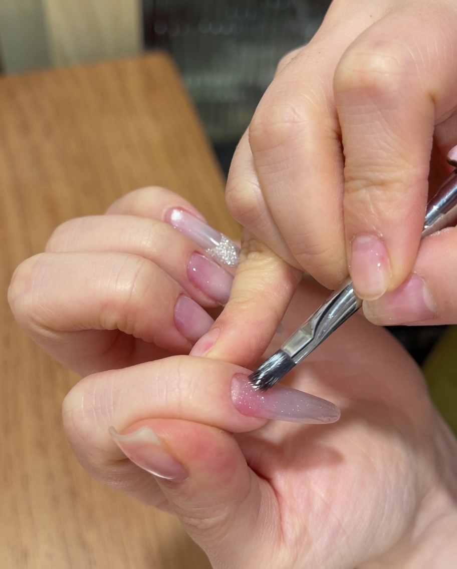 Una mano usa un pennello da unghie per applicare un gel cat eye scintillante su ununghia lunga a mandorla con base sfumata rosa.