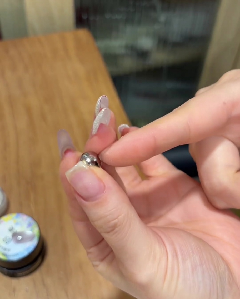 Una mano tiene un piccolo magnete sferico argentato vicino a ununghia dipinta con gel cat eye brillante non polimerizzato.