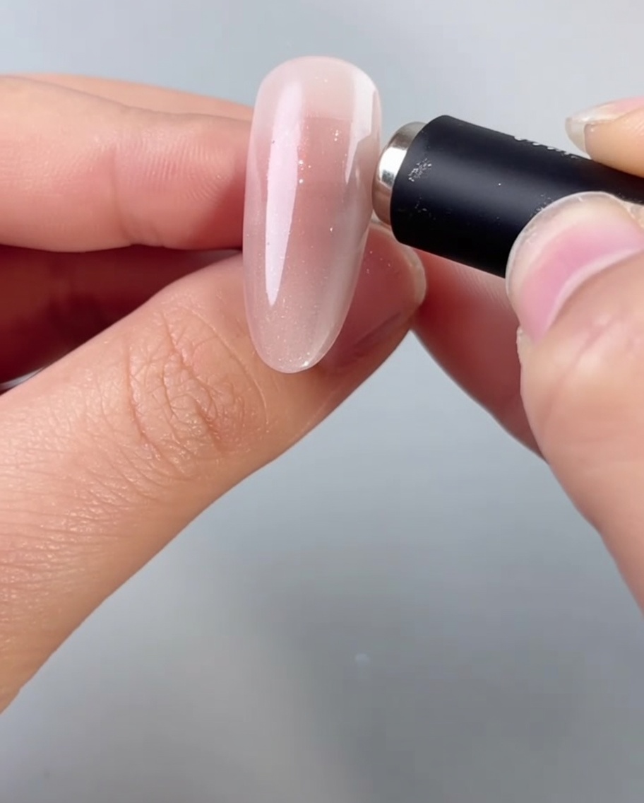 Una mano avvicina un magnete cilindrico a ununghia a mandorla, attivando le particelle brillanti nel gel cat-eye ancora bagnato.