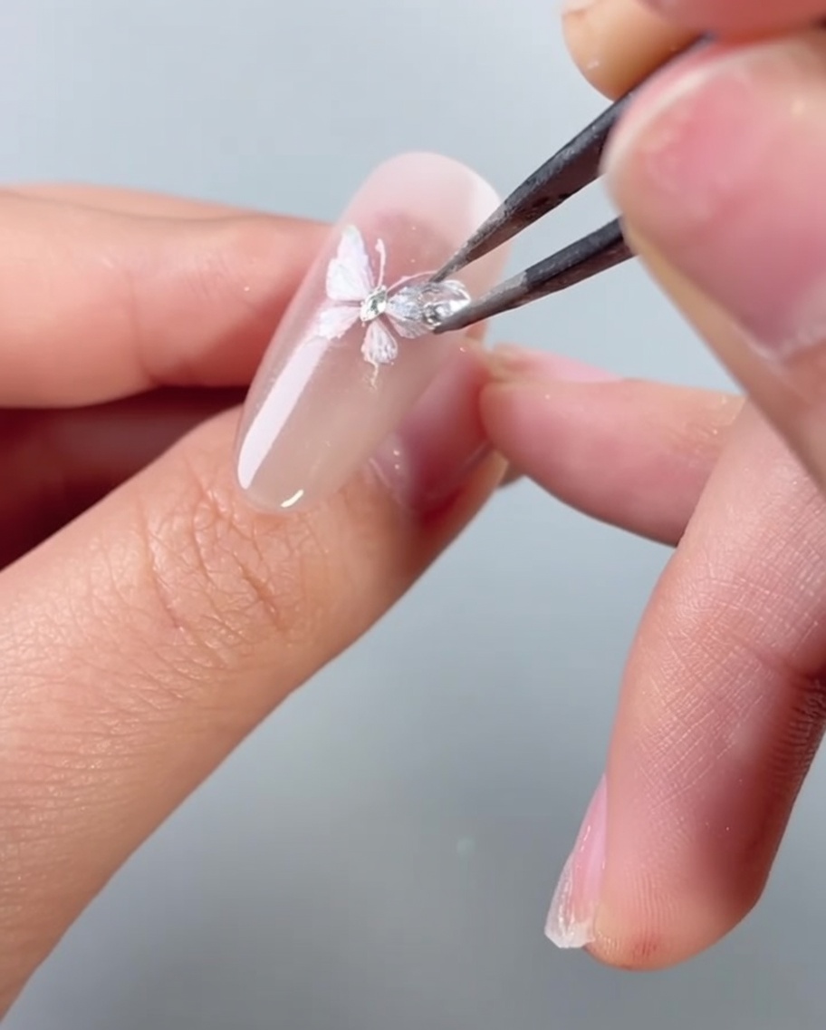 Delle pinzette posizionano un piccolo strass scintillante sopra il design di ununghia decorata con una farfalla.