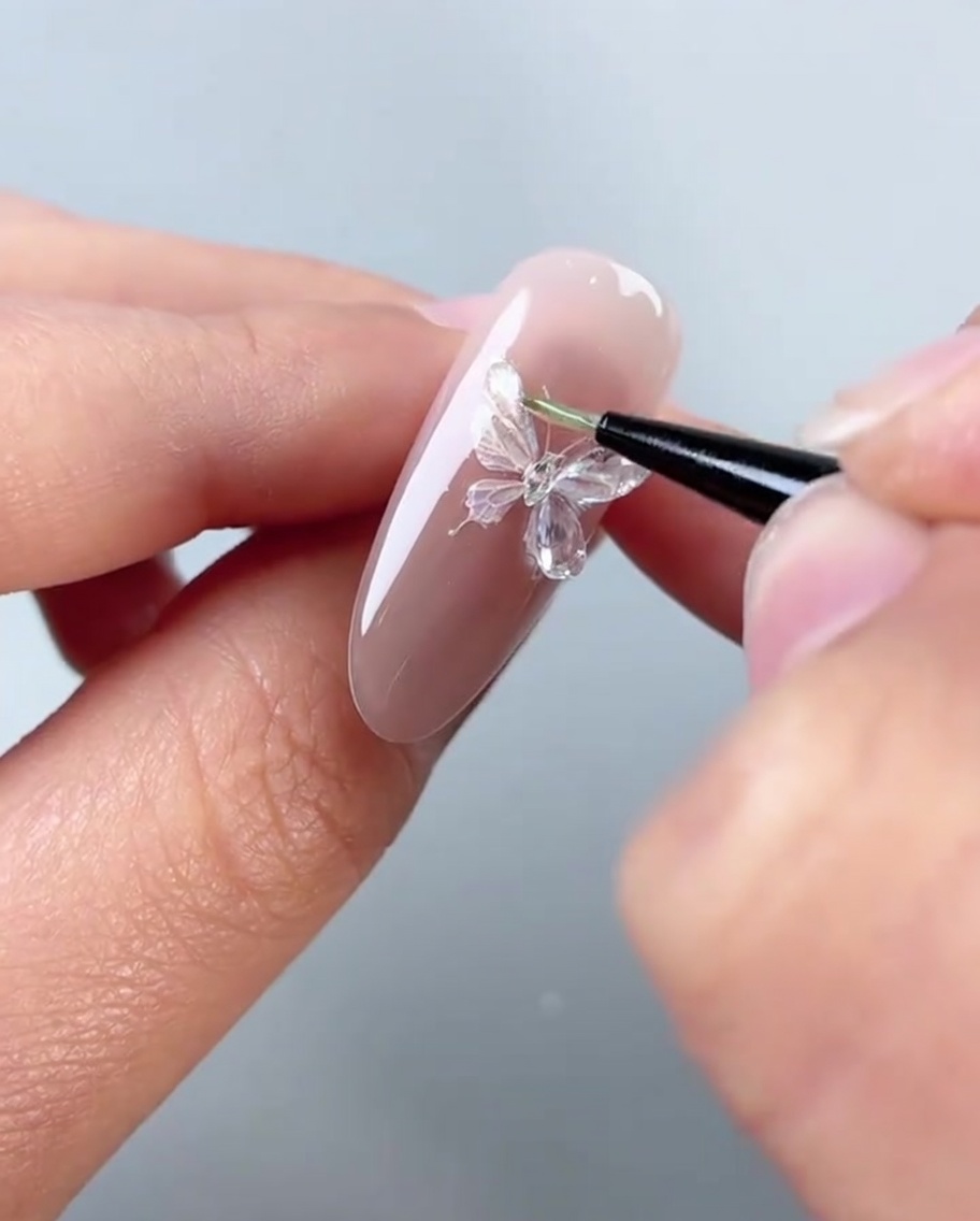 Primo piano di un pennello che applica un gel denso e trasparente per modellare le ali sinistre di un adesivo a farfalla su ununghia rosa.