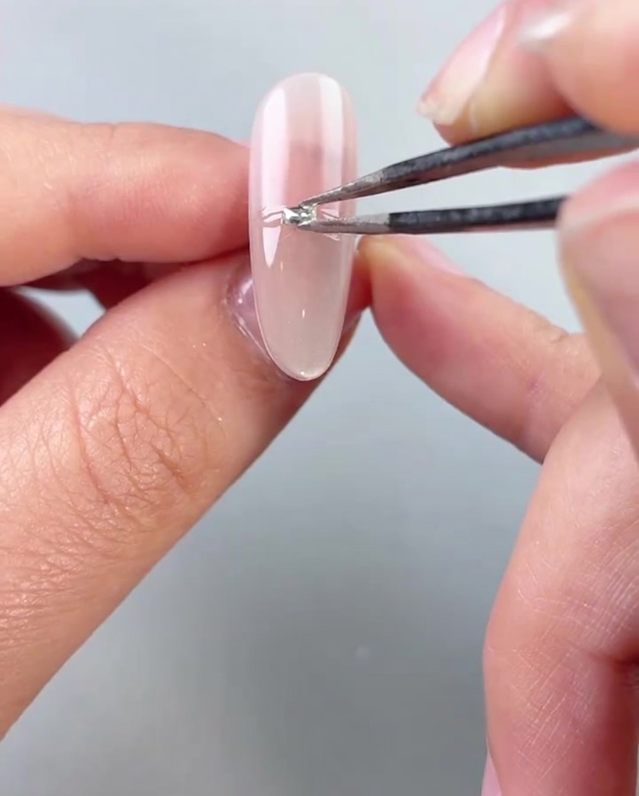 Delle pinzette posizionano piccole perle argentate o strass orizzontalmente attraverso il centro di ununghia laccata col gel.