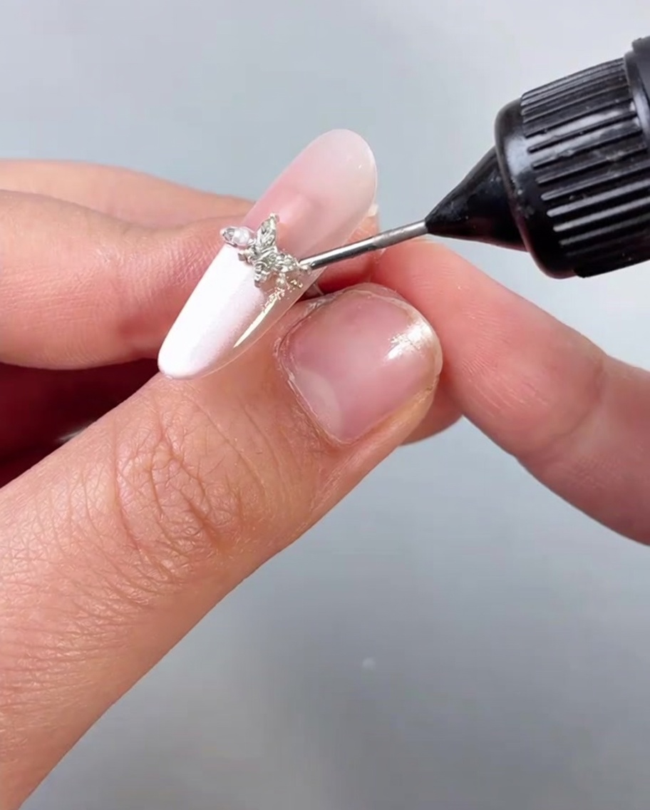 Applicazione di un top coat trasparente o sigillante sopra ununghia impreziosita da un ciondolo a farfalla.