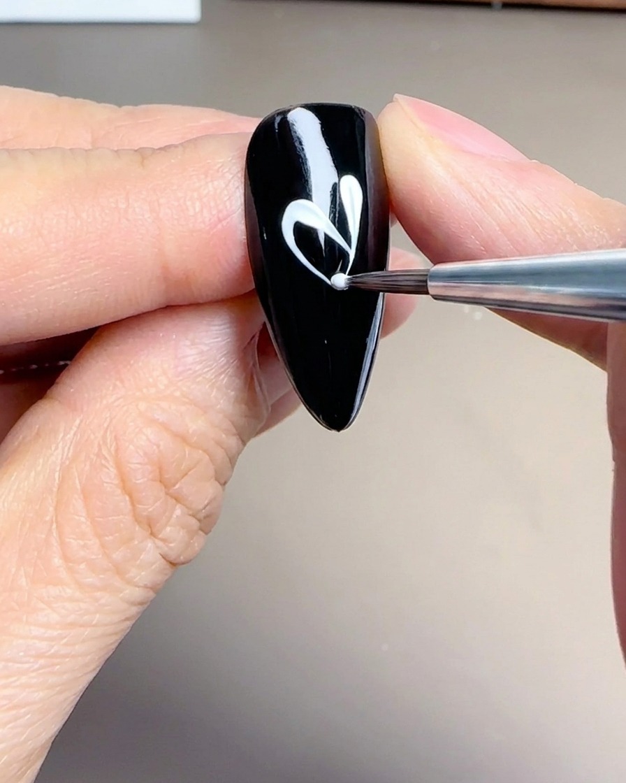 Primo piano di un pennello liner fine che crea forme delicate e allungate di petali bianchi su ununghia nera.