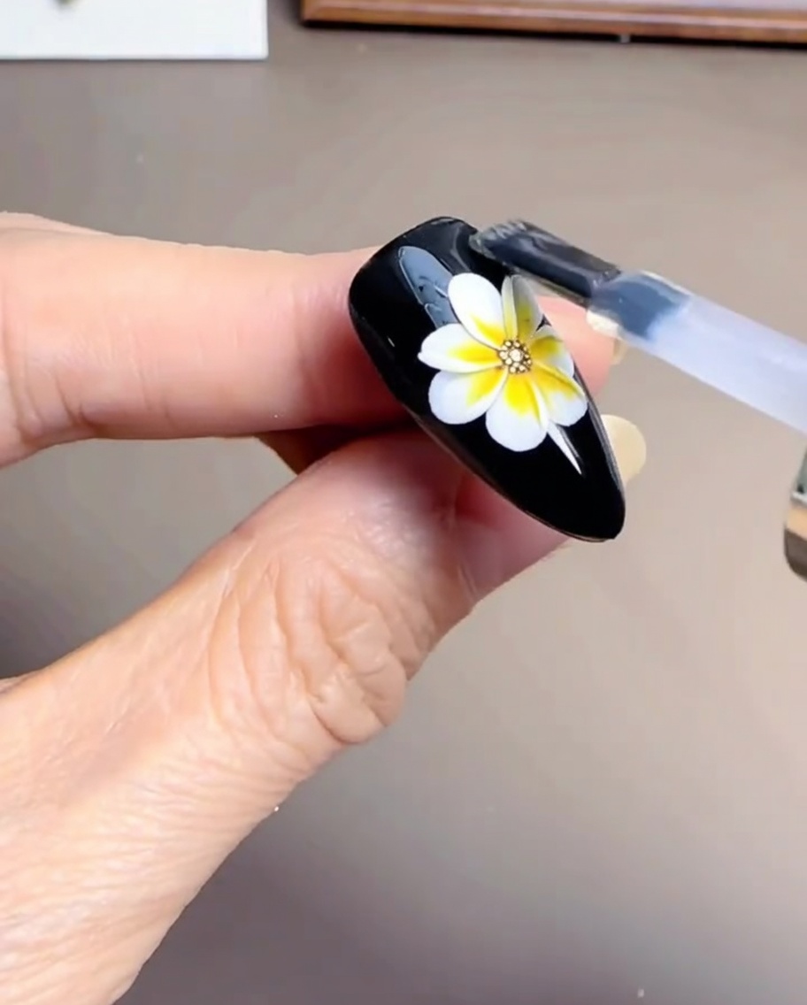 Un pennello per unghie che applica un top coat in gel trasparente e lucido su un design a margherita parzialmente finito su ununghia nera.