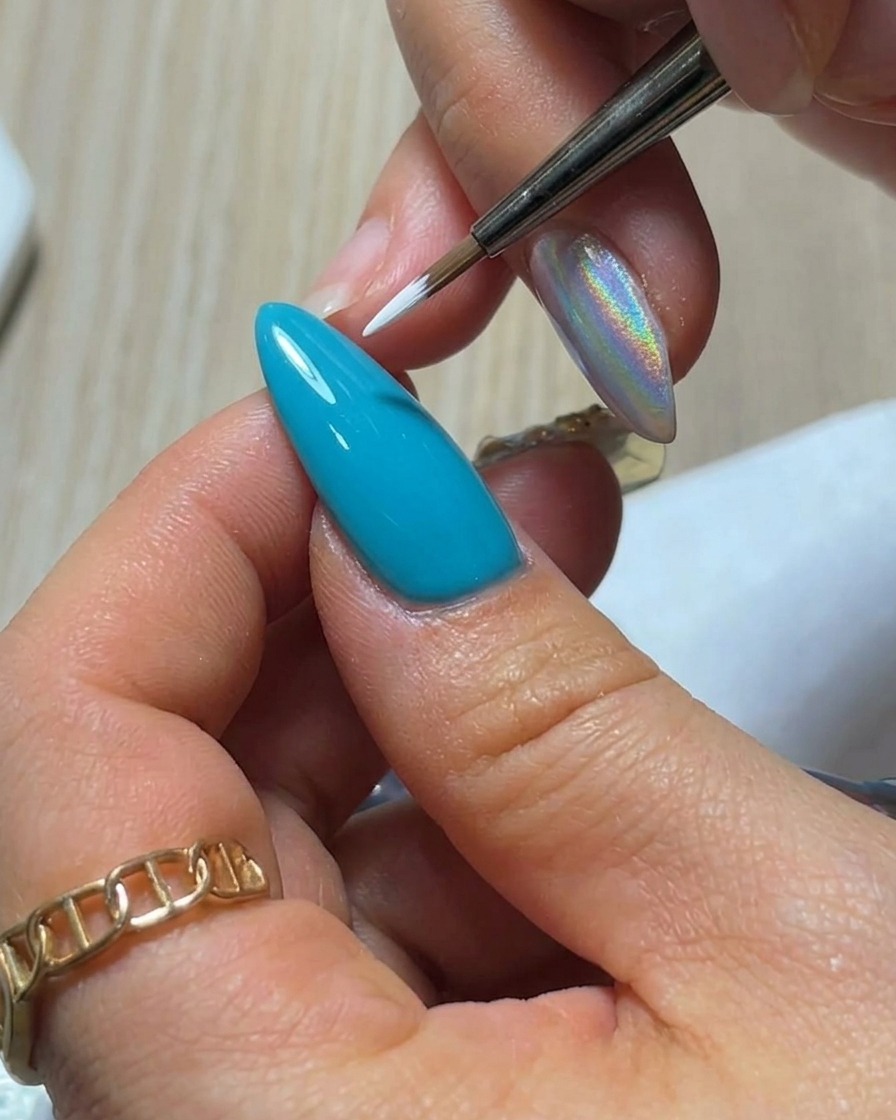 Vista ravvicinata di un pennello liner sottile che applica un singolo petalo di gel bianco a forma di goccia su una superficie ungueale blu brillante.