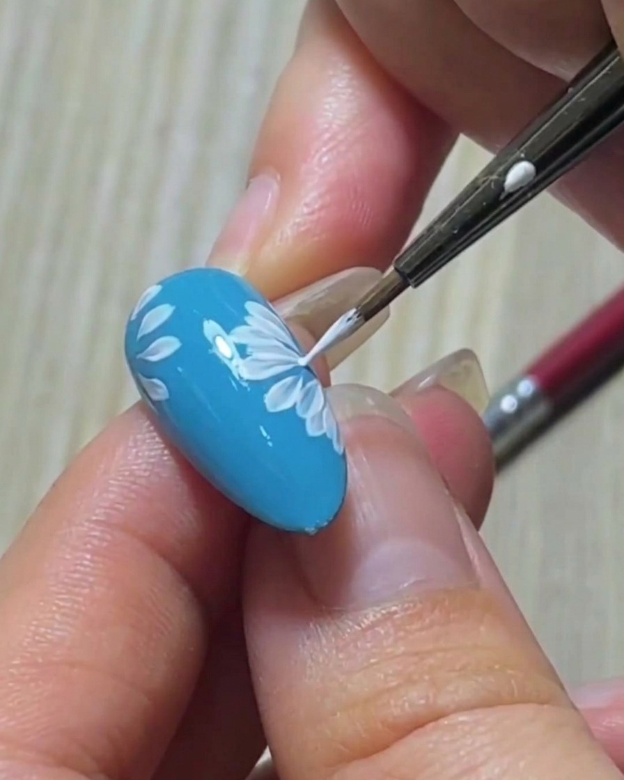 Dettaglio di un pennello liner che rifinisce la punta affilata di un petalo bianco su una base per unghie blu.