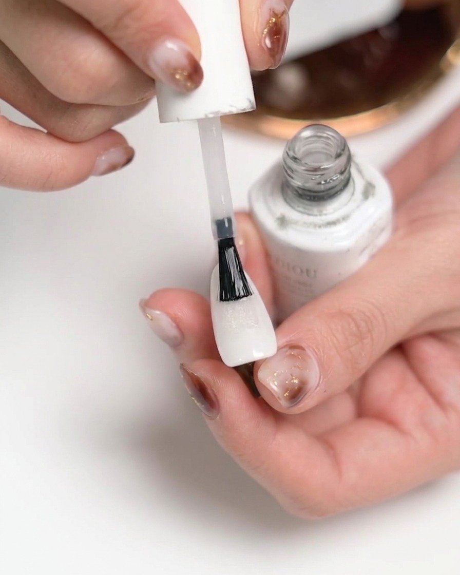 Un primo piano di una mano che applica un top coat opaco trasparente su ununghia dipinta di bianco utilizzando il pennello di un flacone di smalto bianco.