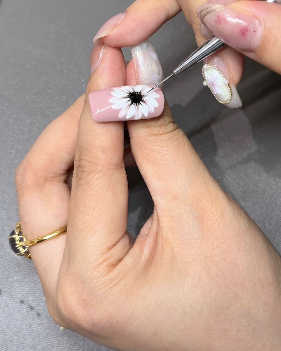 Un pennello fine per dettagli che rifinisce il centro nero scuro di una margherita bianca dipinta su ununghia rosa chiaro.