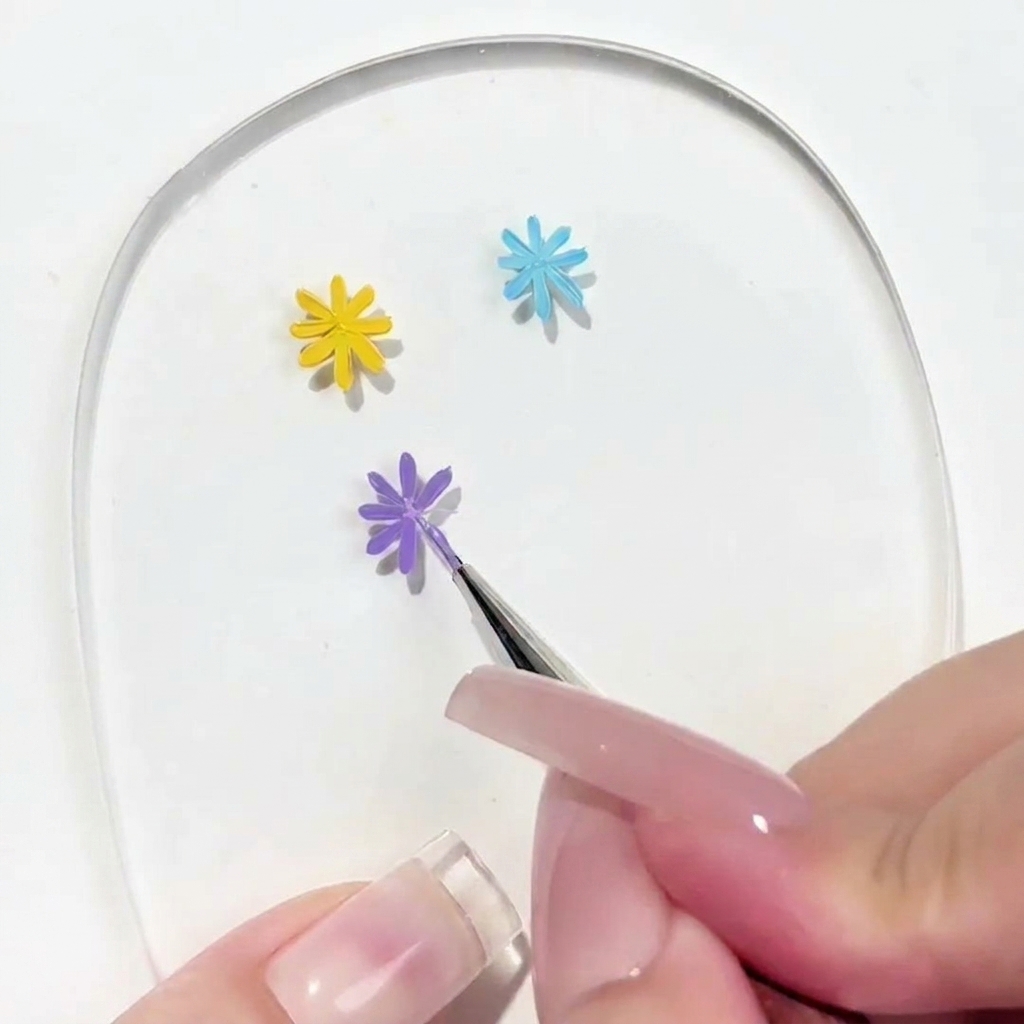 Una vista ravvicinata di un pennello di dettaglio che applica un petalo su una margherita viola parzialmente completata su una tavolozza per nail art trasparente.