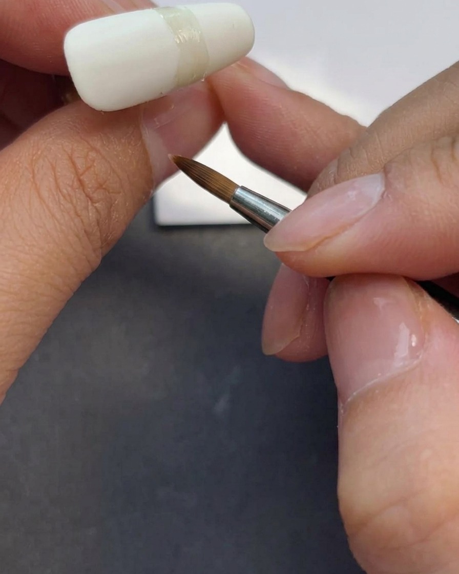 Unonicotecnica usa un pennello da gel per applicare uno strato di top coat opaco trasparente sopra ununghia color bianco lattiginoso.