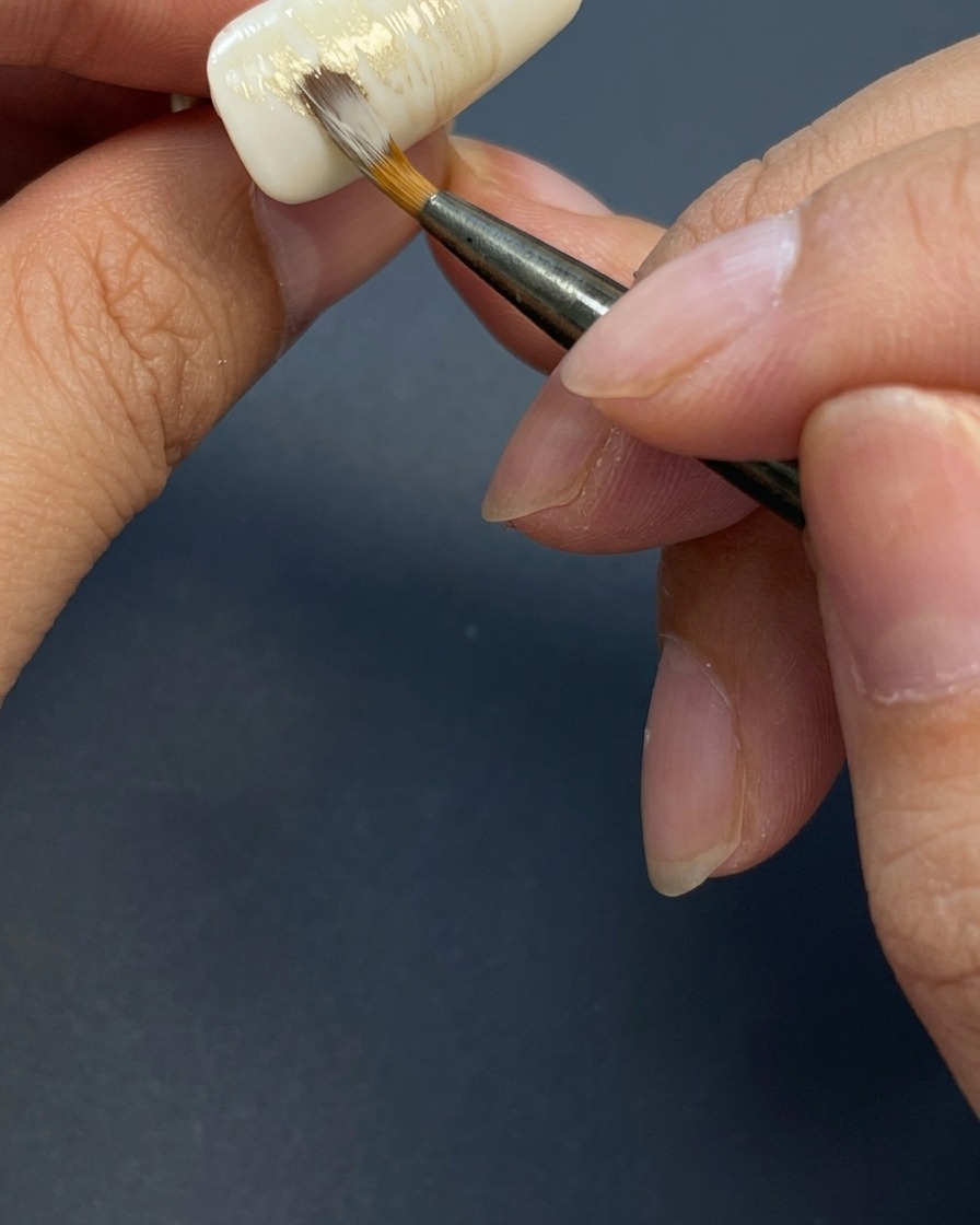 Un pennello applica leggere pennellate orizzontali di un mix di gel color terra su ununghia bianca per creare uno sfondo strutturato.