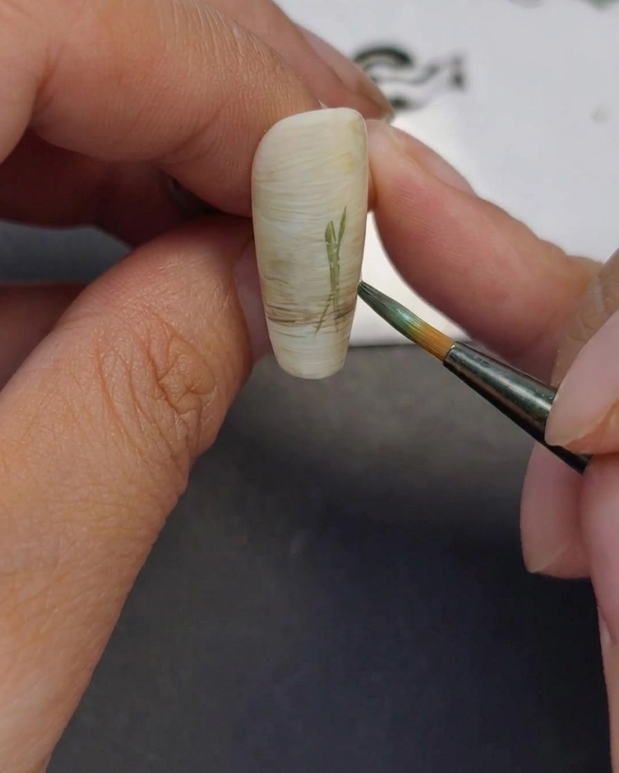 Primo piano di un pennello liner sottile che disegna linee verdi sottili su ununghia per rappresentare gli steli dei fiori.