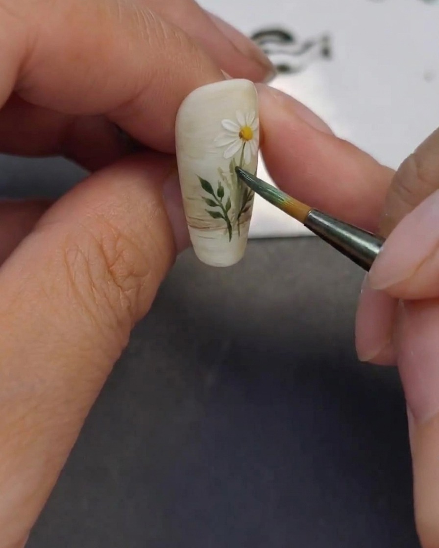 Primo piano che mostra un pennello sottile che aggiunge piccole foglie verdi agli steli dei fiori dipinti sullunghia.