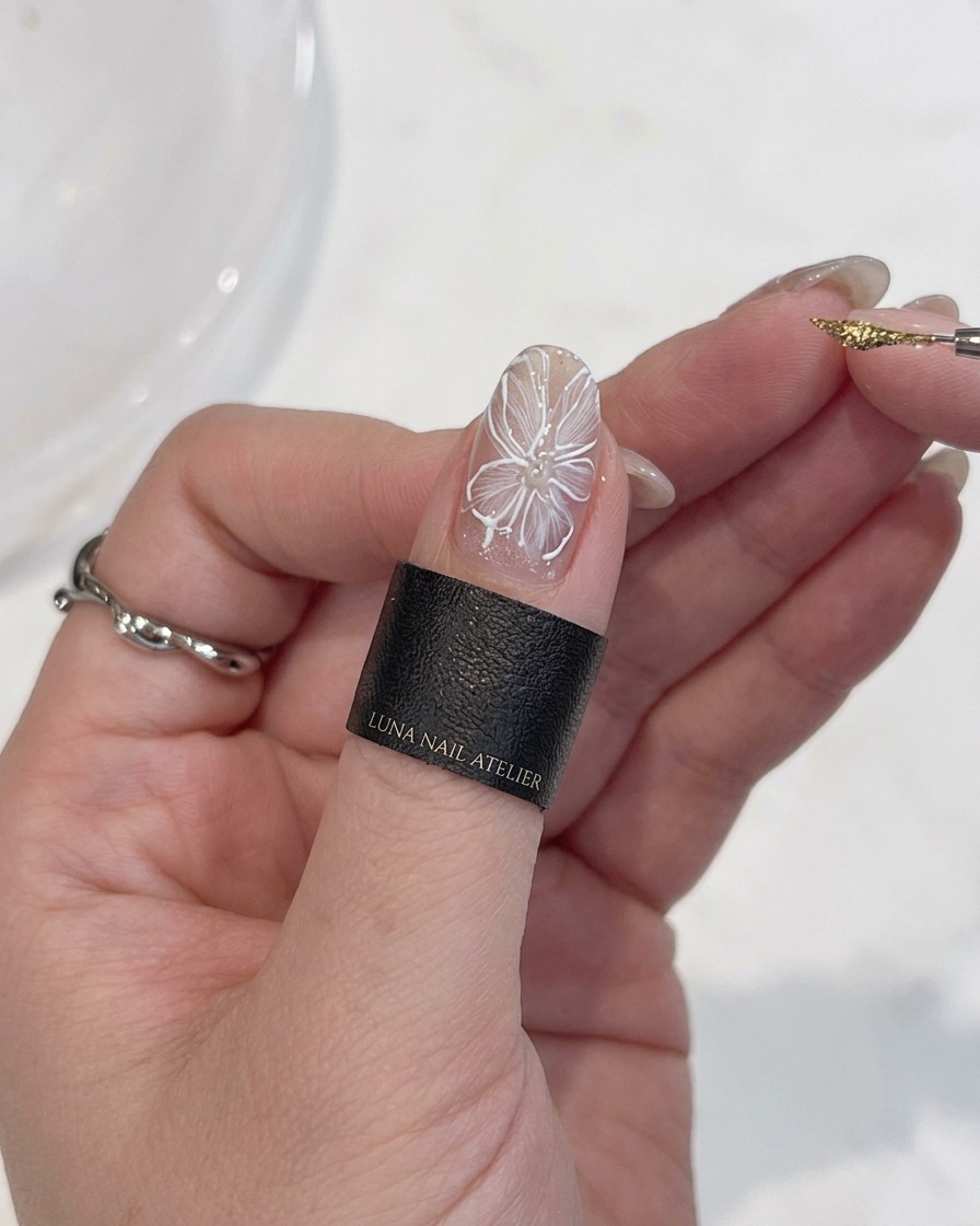 Vista ravvicinata di una mano che tiene un pennello di precisione, applicando un punto preciso di gel metallizzato oro al centro di un fiore bianco dipinto su ununghia.