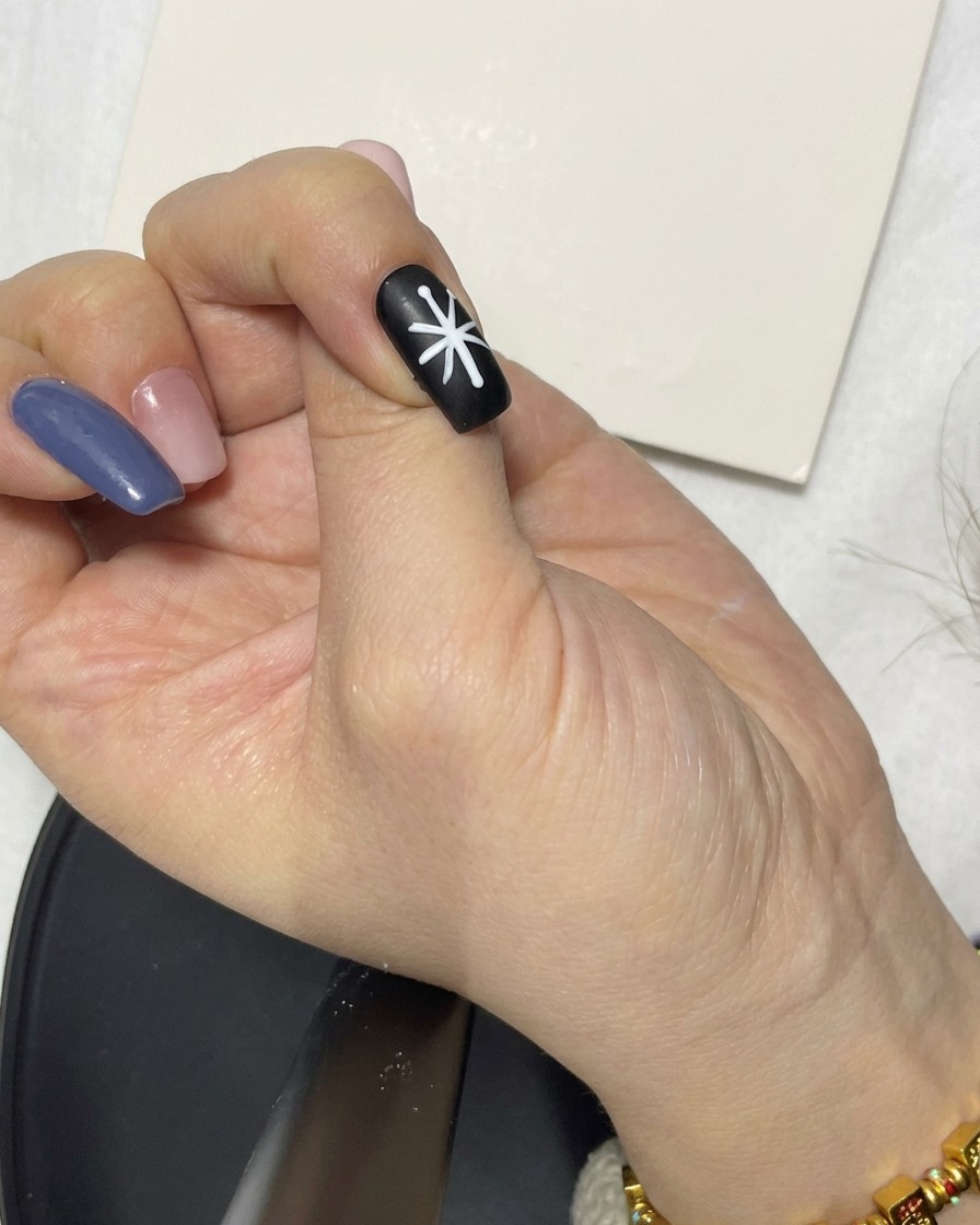 Una mano che usa un pennello liner per rifinire i segmenti di linea bianchi che formano una forma di fiore su ununghia nera.