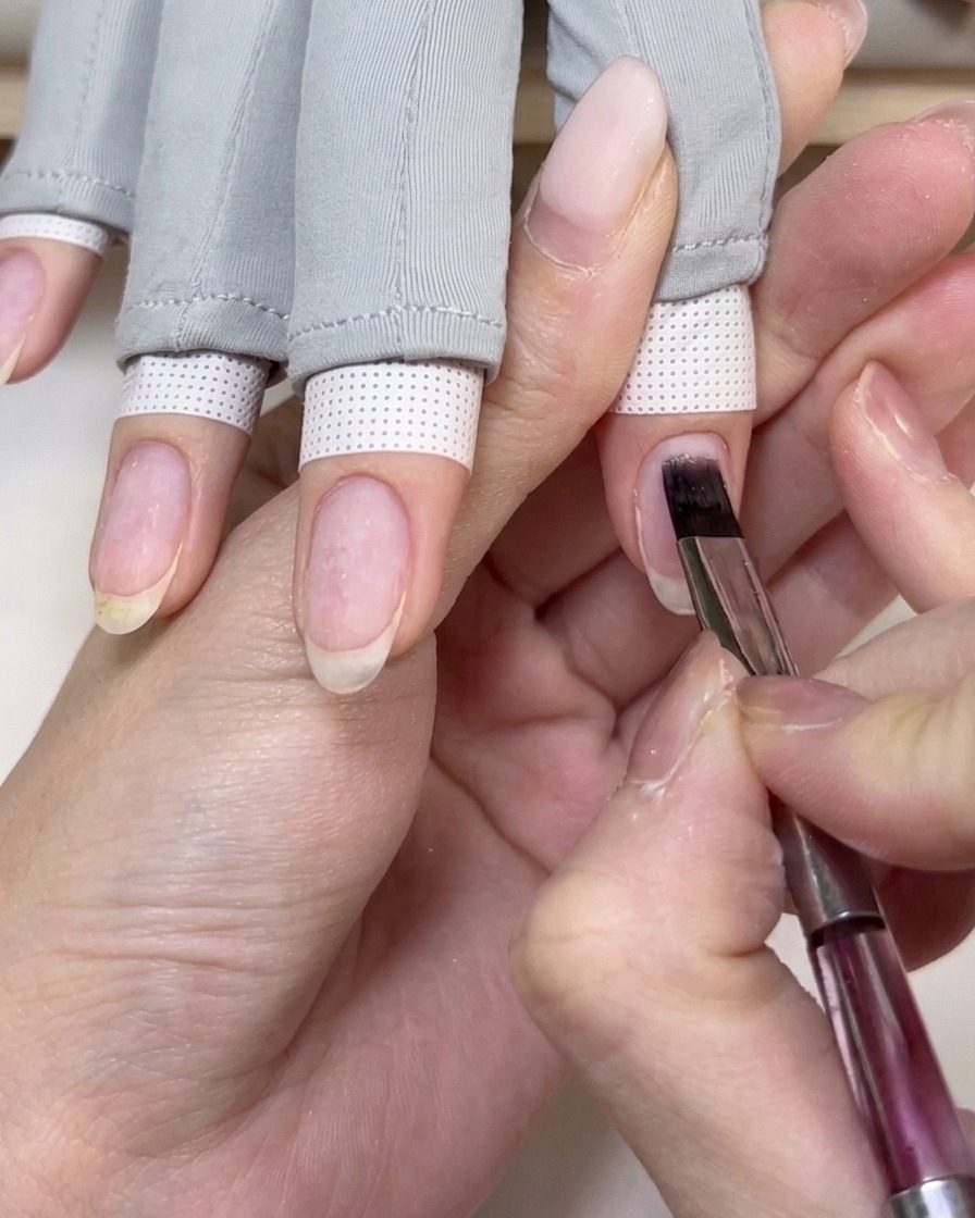 Un pennello piatto per gel che stende uno strato sottile di base coat trasparente sullunghia.