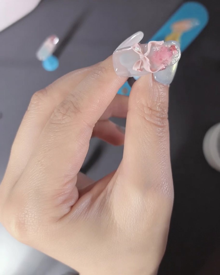 Primo piano di un dito con un charm a fiocco 3D rosa e perle sullunghia.