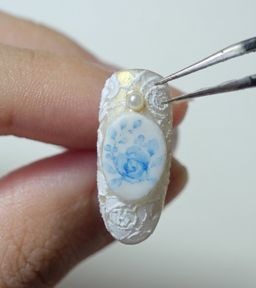 Pinzette che tengono una piccola perlina bianca sopra ununghia con una rosa blu e pizzo bianco in rilievo.