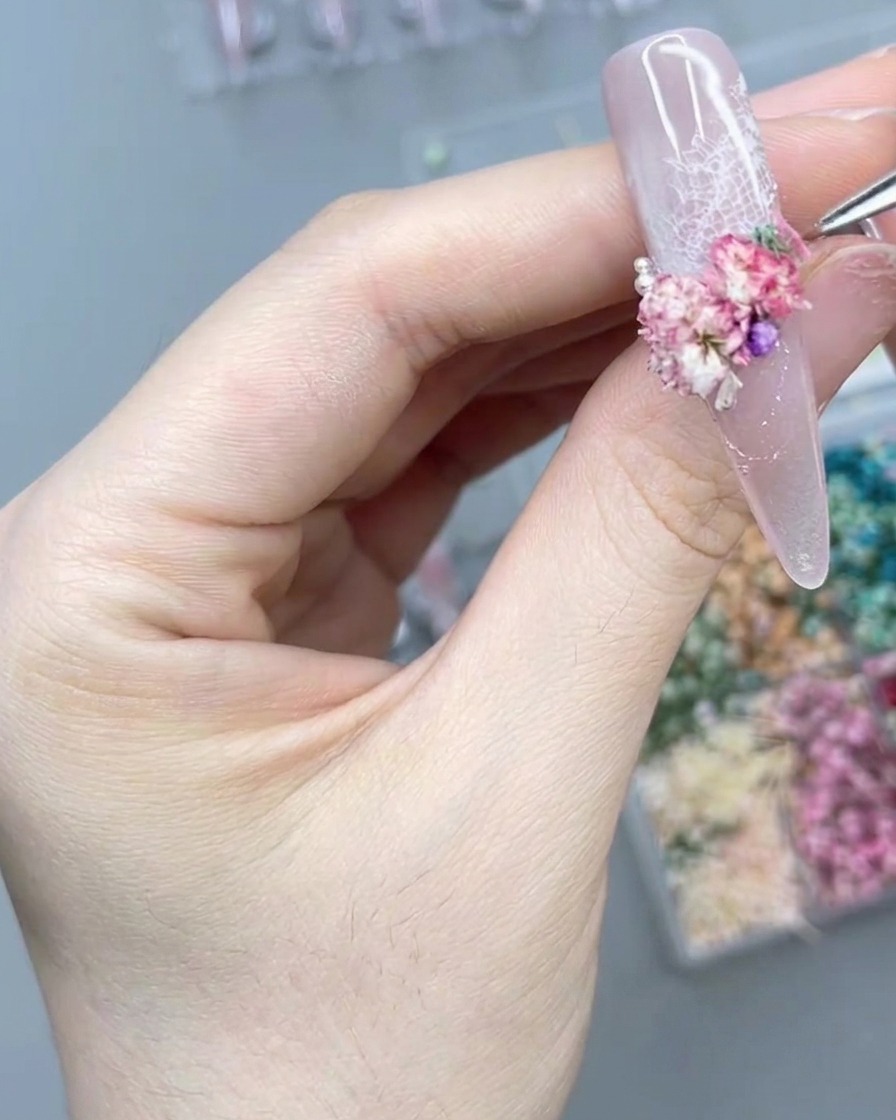 Primo piano di una mano che usa uno strumento di precisione per posizionare una perla vicino ai fiori su ununghia.