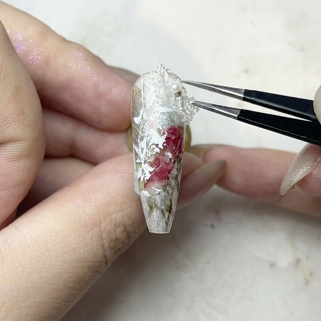 Un paio di pinzette posizionano con cura un piccolo charm a fiore 3D bianco su un punto di colla gel trasparente.