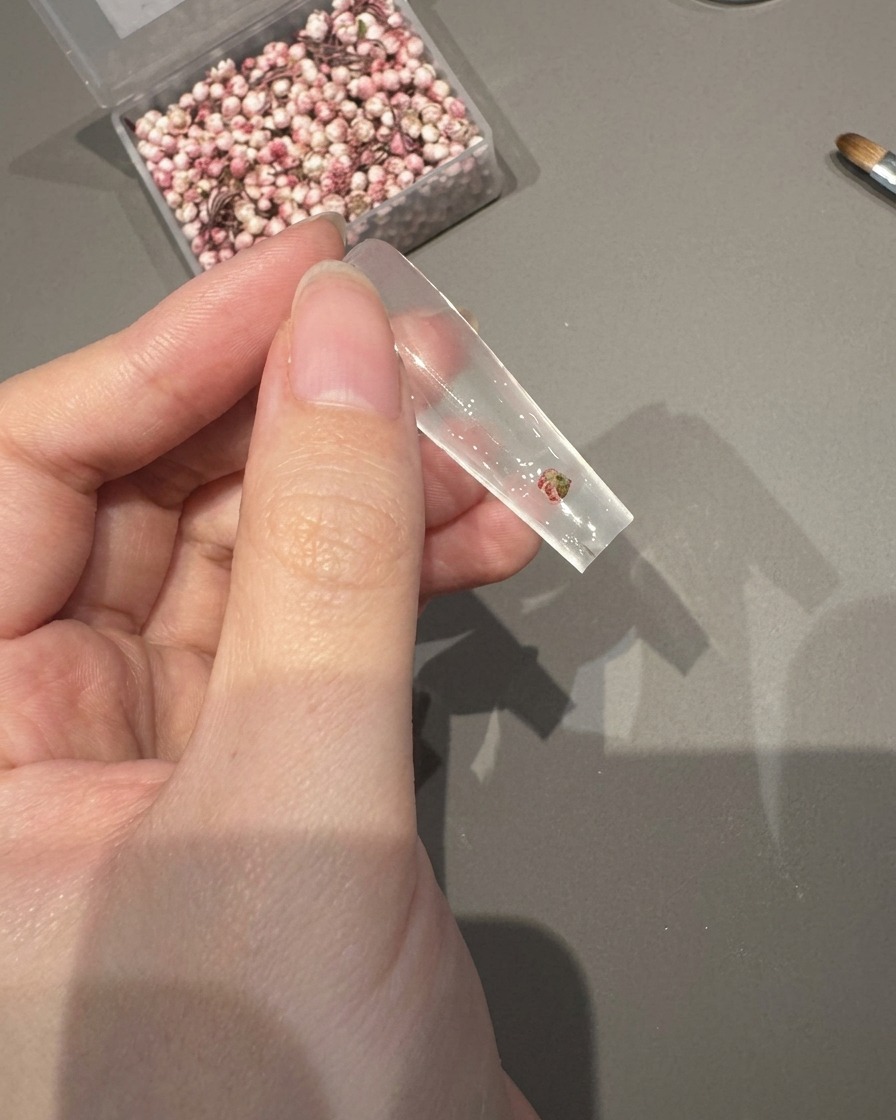 Pinzette che posizionano un piccolo fiore essiccato su una tip trasparente ricoperta di gel adesivo umido.