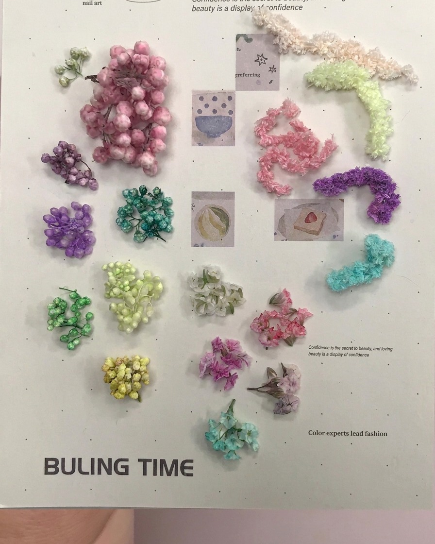Un paio di pinzette dorate che prelevano piccoli fiori sferici viola secchi da una superficie piana.