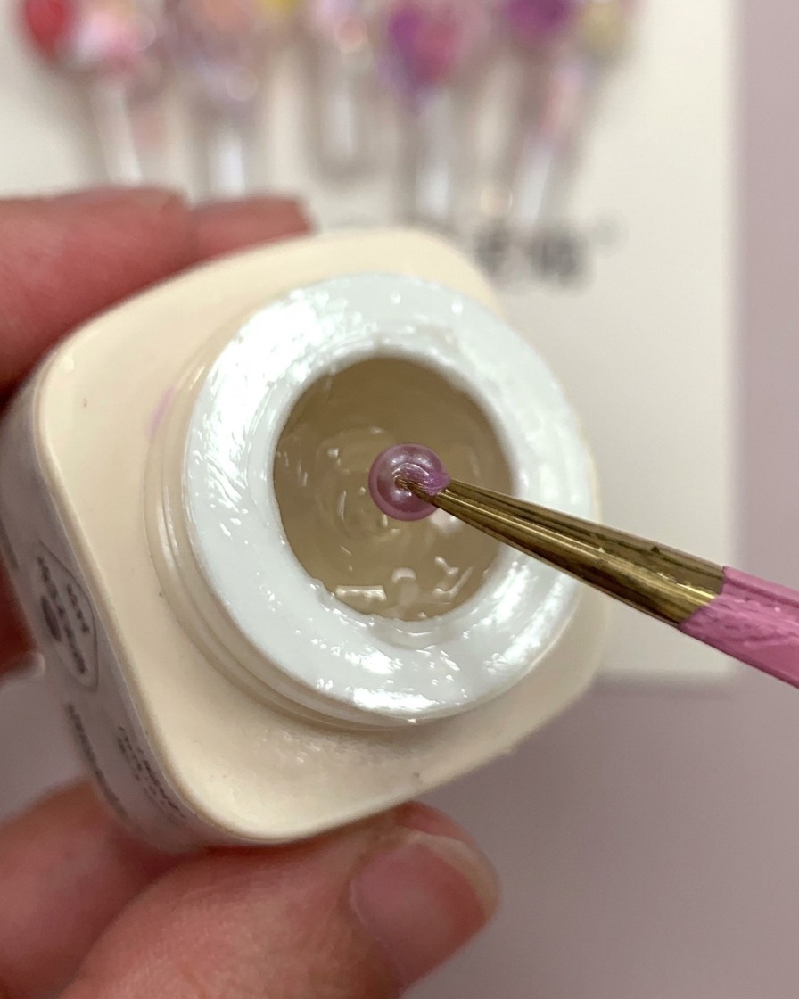 Una mano che tiene una tip con una goccia di gel opaco e fiori sferici viola che vengono posizionati al suo interno.