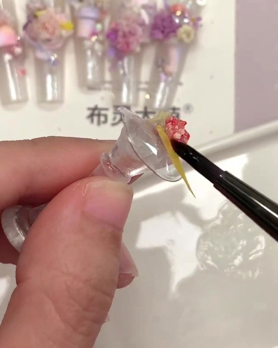 Un pennello fine per unghie che applica del top coat trasparente sotto un piccolo pezzo di fiore secco giallo.