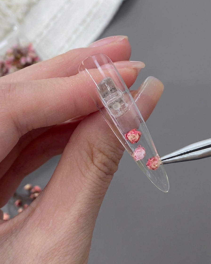 Pinzette a punta fine posizionano un piccolo fiore rosa essiccato accanto a una rosa centrale più grande allinterno di una tip a stiletto trasparente.