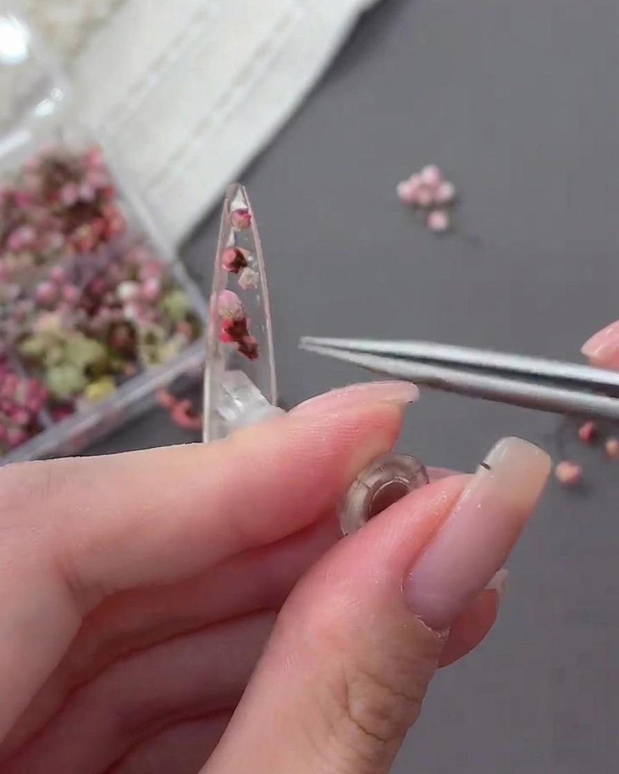 Un primo piano di pinzette che posizionano un minuscolo strass trasparente sulla superficie di una tip non ancora polimerizzata contenente fiori essiccati.