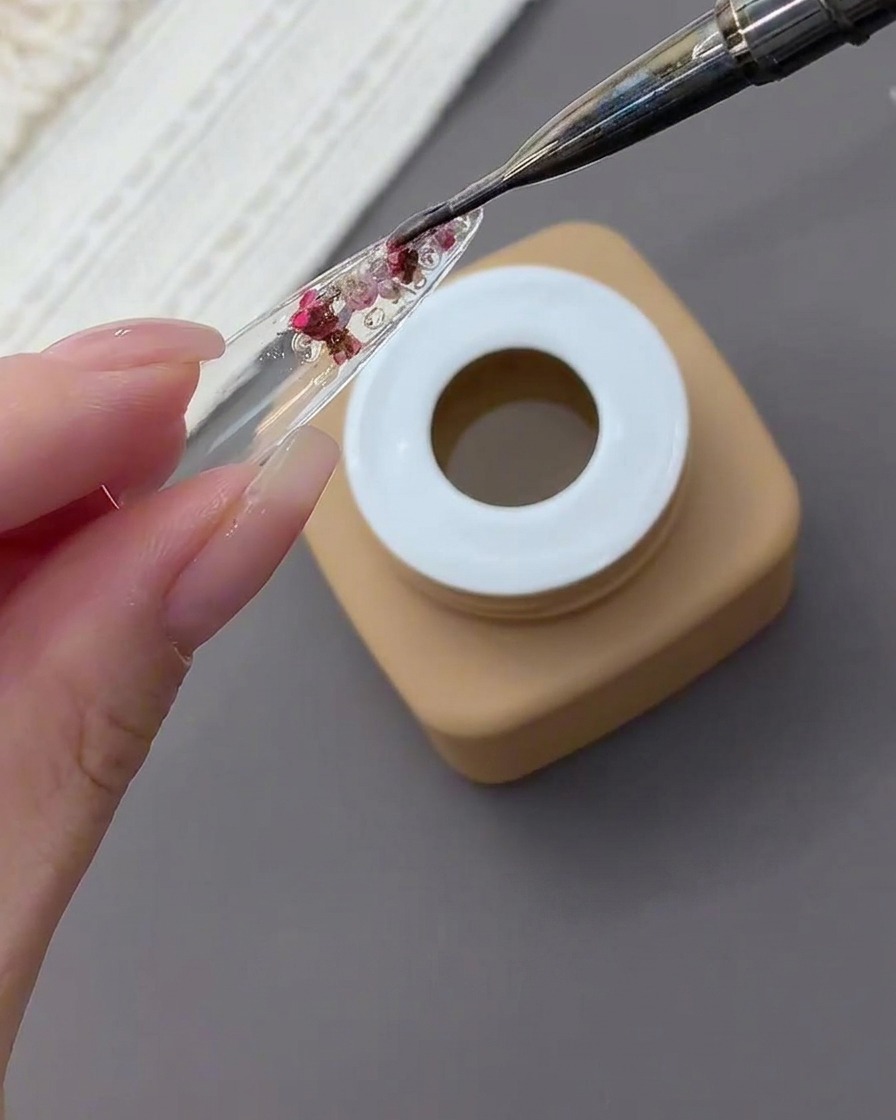 Un pennello da nail art applica uno strato spesso di gel trasparente sopra fiori essiccati e strass incapsulati nel retro di una tip.