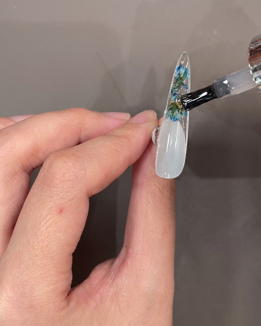 Un pennello applica un top coat trasparente sopra fiori e pietre sul retro di una punta.