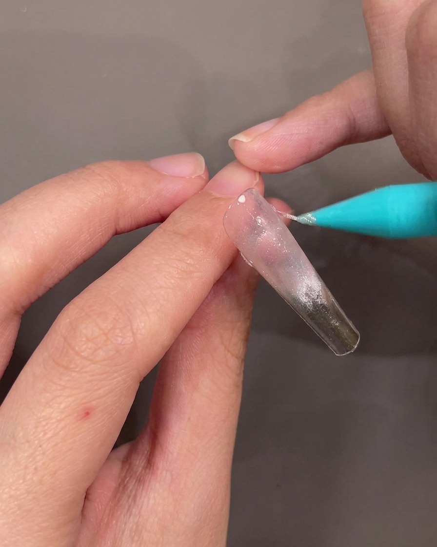 Dettaglio di una mano che applica una sottile linea diagonale di gel glitterato argentato su una punta trasparente.