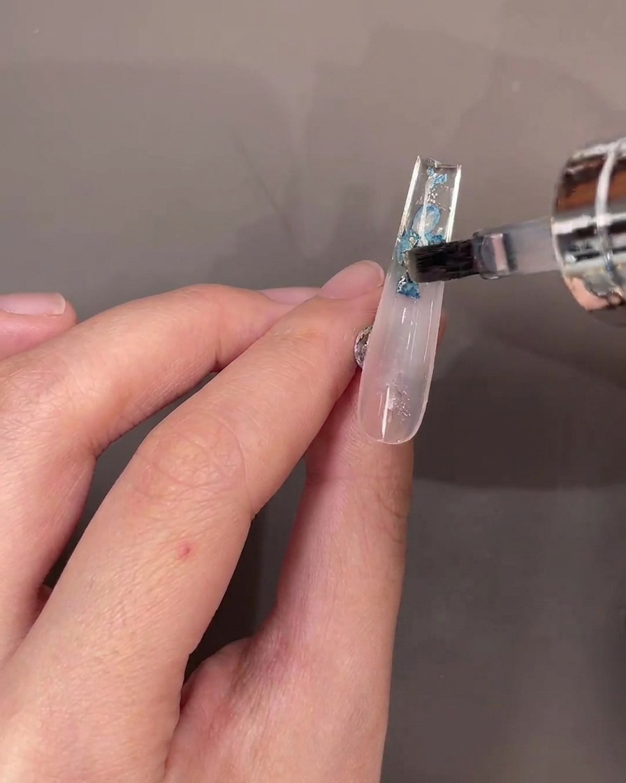 Un pennello applica uno strato di top coat trasparente sopra i fiori secchi sul retro di ununghia trasparente.
