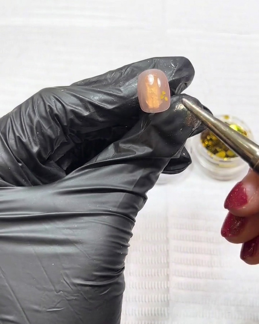 Un primo piano di ununghia che mostra glitter oro e argento posizionati con uno strumento su una base gel nude.
