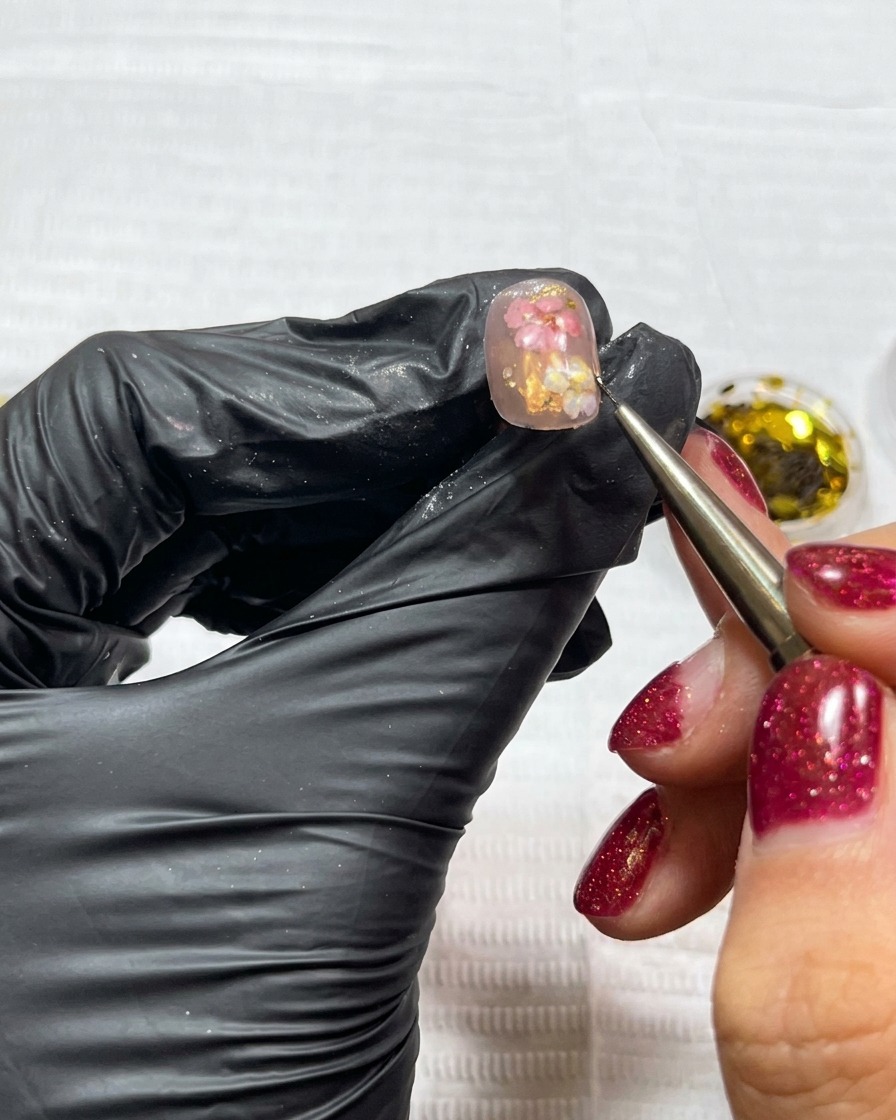 Un pennello di precisione che applica piccoli frammenti irregolari di foglia oro su ununghia già decorata con fiori secchi.
