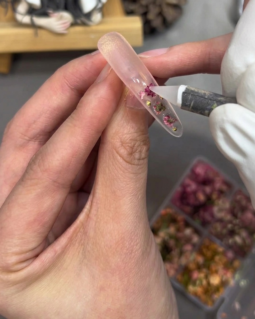 Dettaglio di uno strumento che posiziona piccole perle dorate e strass rosa su una tip ricoperta di gel trasparente.