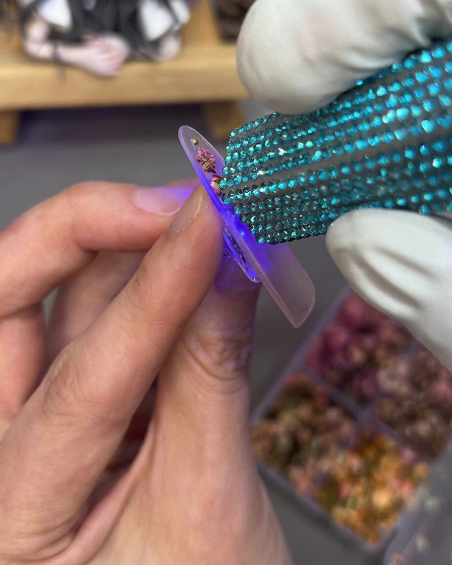 Una mano tiene una mini lampada UV su una tip che presenta fiori secchi e strass sulla superficie.