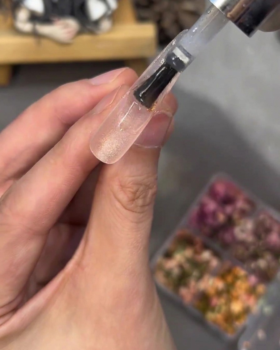 Un pennello applica uno strato spesso di gel trasparente su una tip con fiori e strass, levigando la superficie.
