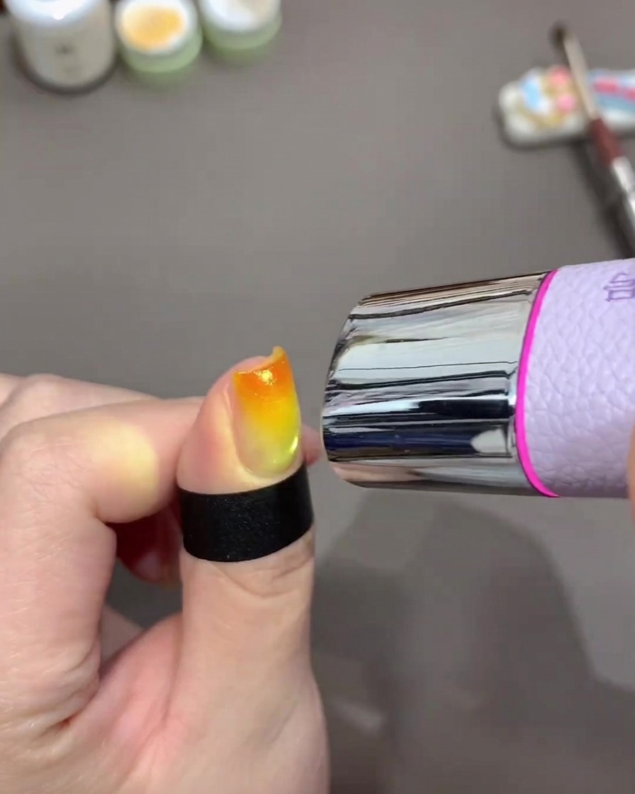 Una mini lampada UV portatile posizionata sopra ununghia per polimerizzare un design gel con gradiente giallo e arancione.