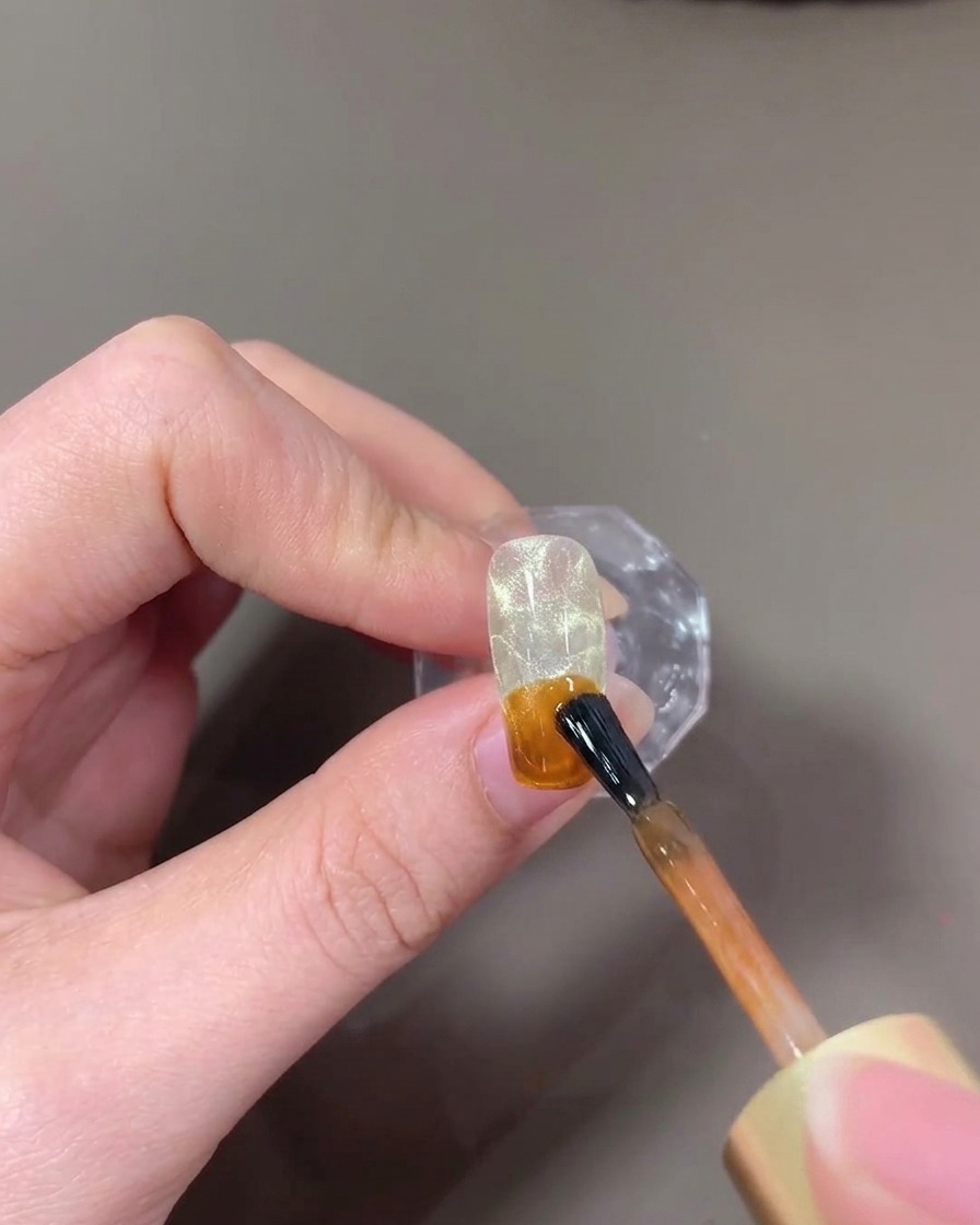 Un pennello che applica un gel traslucido color ambra sulla punta di ununghia.