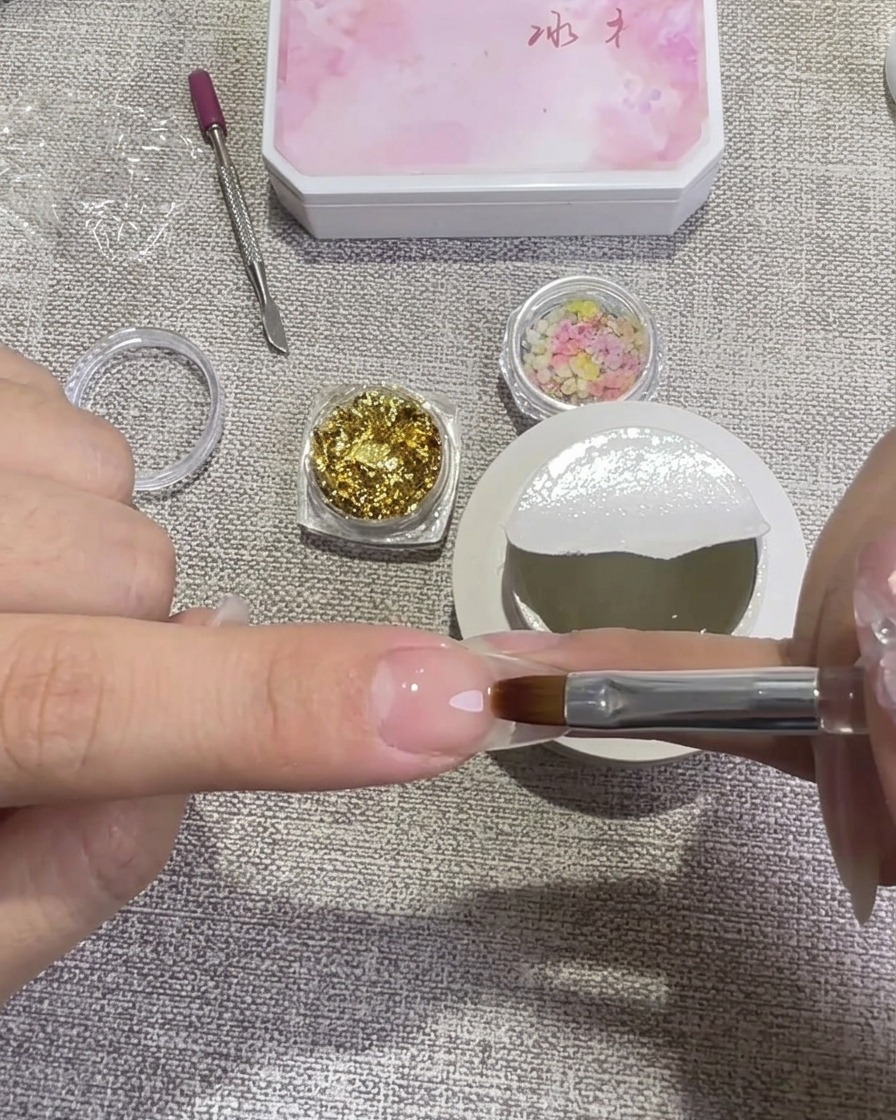 Una mano che utilizza un pennello piatto per applicare uno strato di hard gel trasparente su ununghia preparata, con piccoli barattolini di foglia doro e fiori secchi sullo sfondo.