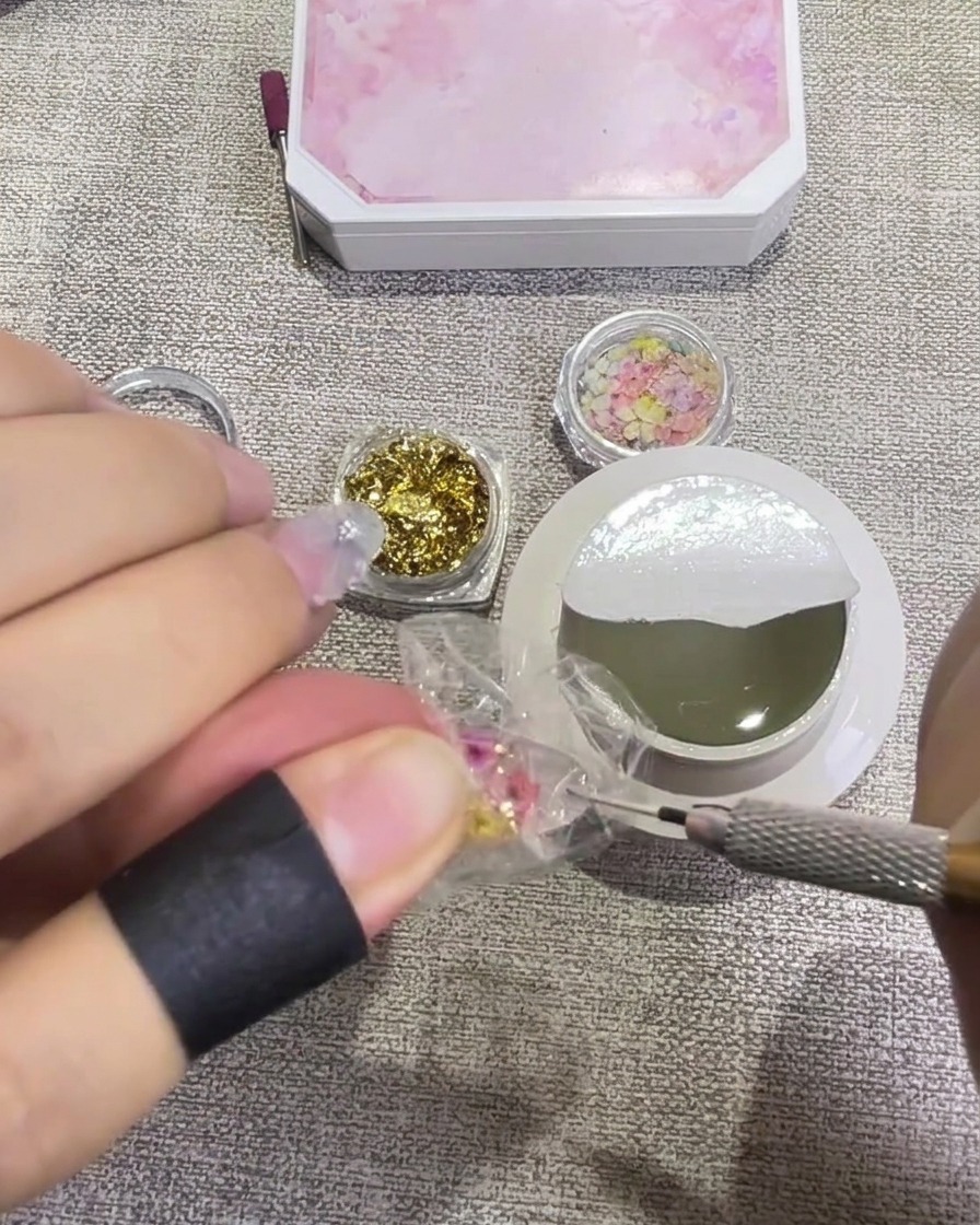 Un pennello applica un top coat trasparente e lucido sopra ununghia decorata con fiori pressati e foglia doro, conferendole un aspetto bagnato e traslucido.