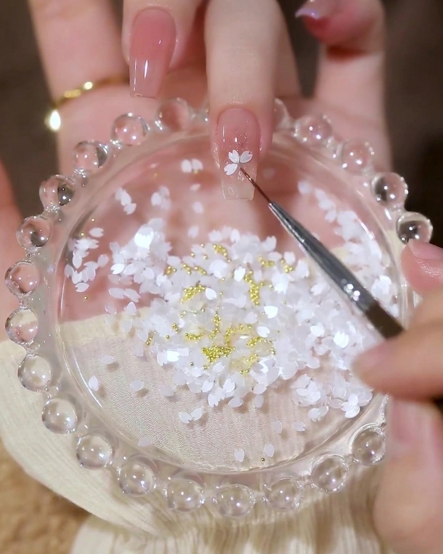 Primo piano di un pennellino di precisione che posiziona paillettes bianche a forma di petalo sullunghia creando un fiore.