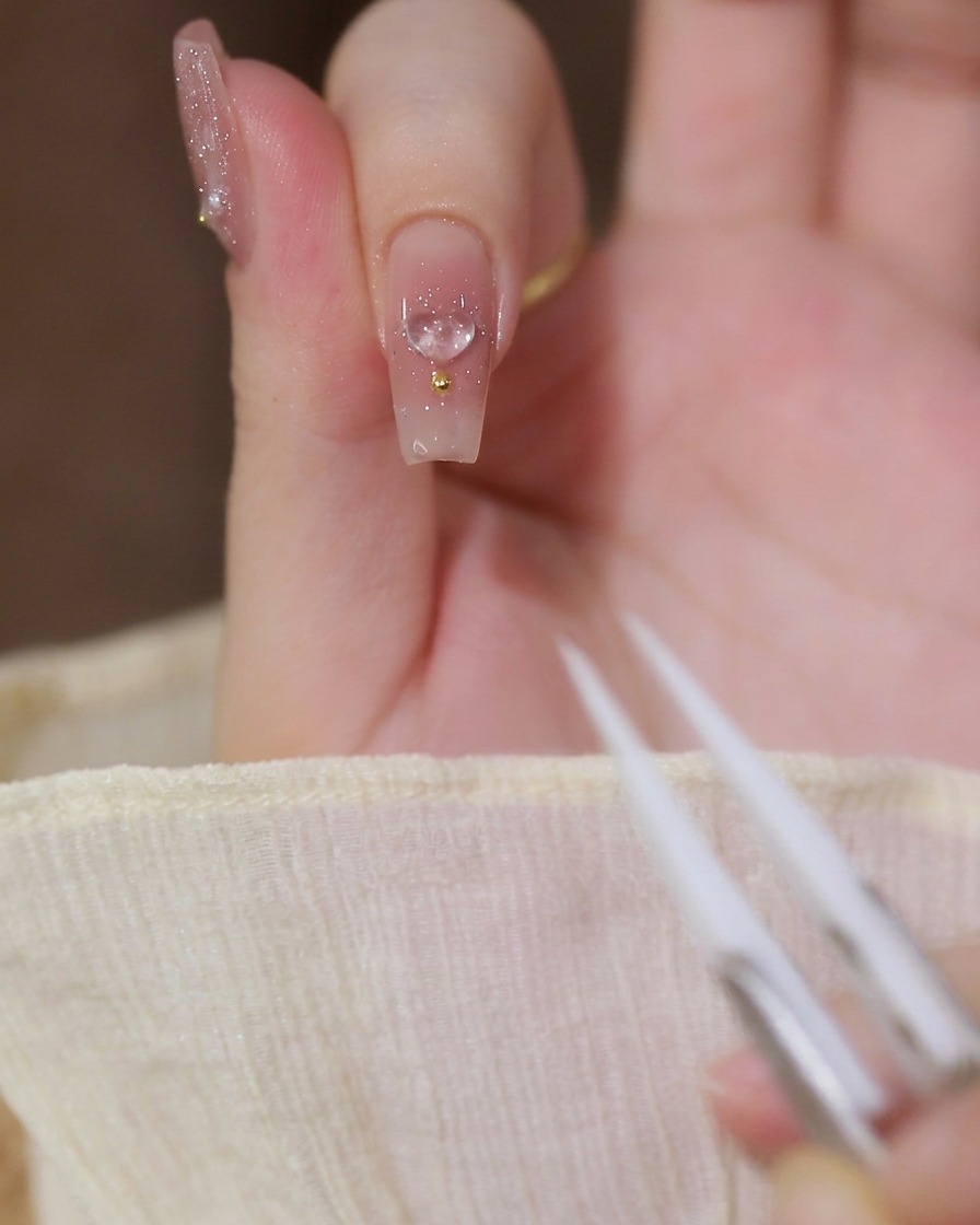 Primo piano di un dito indice con una base in gel nude, decorato con un charm a cuore 3D trasparente e una perlina dorata, con pinzette visibili in primo piano.