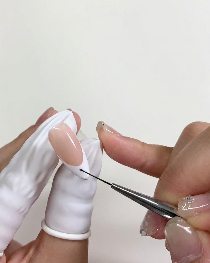 Un pennellino per nail art posiziona un minuscolo punto di smalto gel nero sulla punta bianca di ununghia, sopra uno strato di blooming gel non polimerizzato.
